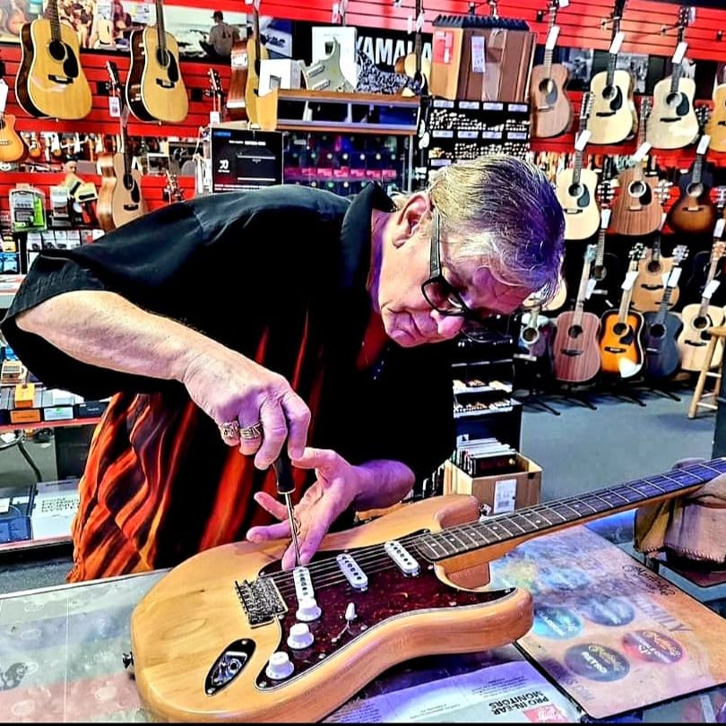 A person carefully adjusts a wooden electric guitar with a screwdriver in a vibrant guitar store. Numerous acoustic guitars hang on a red wall.
